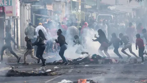 Getty Images Women and children run across a road. There is smoke billowing across the road and there is rubble, debris or rubbish strewn across.