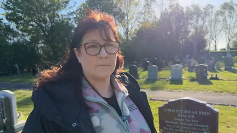 Lyndsey Wood is wearing a black coat and is standing next to a headstone. Behind her is rows of other headstones in the cemetery.
