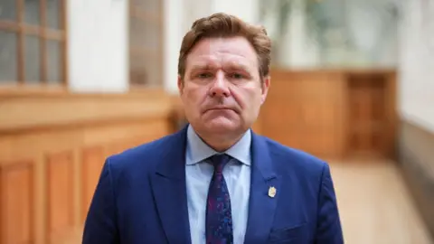 PA Media A man, John Cotton, looks into the camera. He has short reddish hair and is wearing a dark blue jacked, with a blue-patterned tie and a light blue shirt. Behind him is a lot of wooden pannelling.