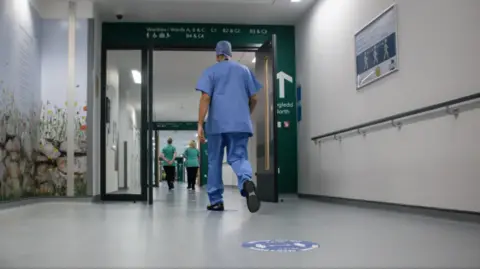 A medical professional wearing blue scrubs walking down a hospital corridor 