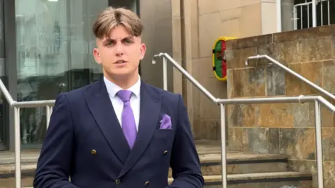 A man, George Finch, has short brown hair and is wearing a white shirt, a blue suit and purple tie. He is standing in front of steps and metal railing.