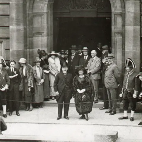 Walsall Archive A black and white image of Pat Collins, in black suit and top hat, standing outside the council house next to a woman also wearing a dark outfit. Many people are gathered behind them.