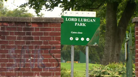 A sign sign that says Lord Lurgan Park. The photo is taken from outside the park. There is graffiti on the brick walls. Grass, bushes and trees are in the background