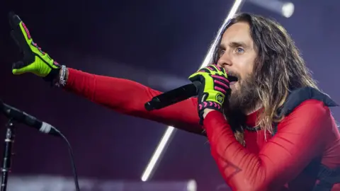 Getty Images Jared Leto stands under concert lighting, singing into a handheld microphone. He has long hair and a beard and is dressed in a red, sheer and fitted top with neon green and pink gloves. One arm is stretched outward toward the audience while the other holds the microphone close to his mouth. Bright white stage lights form diagonal lines in the background. CLARKSTON, MICHIGAN - AUGUST 06: Actor/Singer Jared Leto of Thirty Seconds To Mars performs during their Seasons World Tour 2024 at Pine Knob Music Theatre on August 06, 2024 in Clarkston, Michigan. (Photo by Scott Legato/Getty Images)