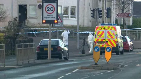 Forensic experts in white overalls are seen near a car and a police van on Drumfrochar Road in Greenock. The area has been cordoned off with police tape