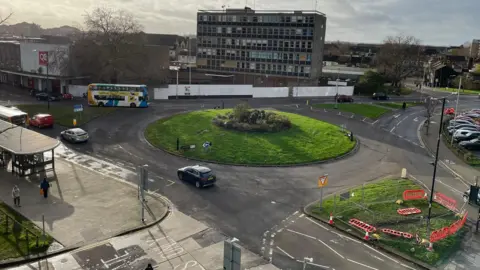 Shot from above of a roundabout covered in grass, with a sculpture in the centre. There are cars going round the roundabout. There is a six-storey building on the far side of the roundabout, and smaller buildings to the left.