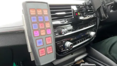 A control panel attached to the dashboard of a car, with brightly colored buttons: orange, blue, pink and red. The buttons all say different thoughts - one says Blues and the other says 999.