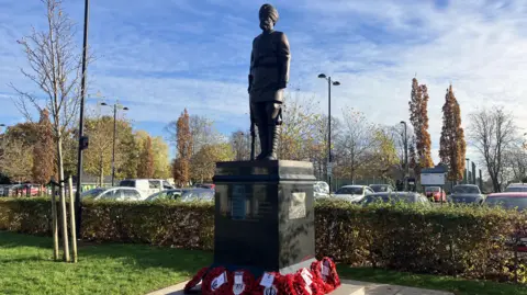 BBC A bronze figure of a male soldier wearing a turban stood on a plinth, which has poppy wreaths placed at its base. There is a green hedge and a car park behind it.