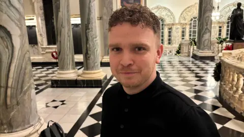 Ryan Murphy. He has short brown curly hair, wearing a black polo top. He is standing inside Belfast City Hall with checkerboard flooring and marble columns in the background.