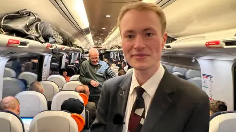 A man wearing a CrossCountry uniform - a dark blazer and tie - is pictured smiling at the camera. He is standing on a packed train. 