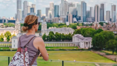 A woman with a backpack looks out over Greenwich Park towards the modern skyline of Canary Wharf in London.