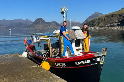 Amy McKinnon Brooke Lamond stands on a small creel fishing boat with James Robertson. She wears a long-sleeve black top with yellow and pink fishing overalls on top. James wears a short-sleeve blue top with blue and yellow fishing overalls. It is a sunny day and you can see the still water around them and hills in the distance.