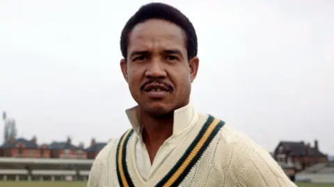 A colour head-and-shoulders shot of a man in his 30s in cricketing attire, a white cricket top with a black-and-yellow stripe. He is standing facing the camera in an empty cricket ground on an overcast say. He has short black hair and a thin black moustache.