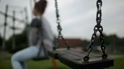 A teenager sits on a swing in a park with her back to the camera. She is wearing jeans and a grey jumper. An empty swing is in the foreground of the image.