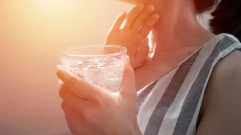 A woman in a stripy top holding a glass of ice water and touching her neck.
