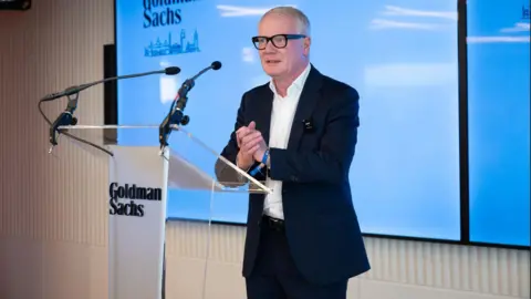 A man wearing black glasses and a dark suit stands in front of a lectern that has Goldman Sachs written on it. He is clasping his hands together. Behind him is a large TV screen