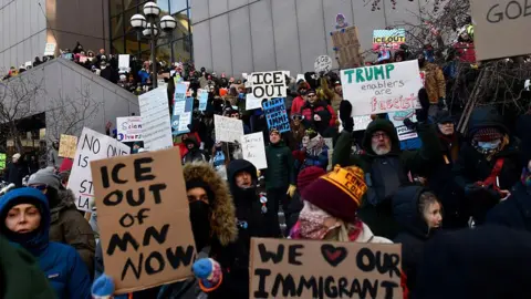 A crowd of protesters waving anti-ICE signs and wearing winter clothes stand on the stairs of a large concrete building in Minnesota