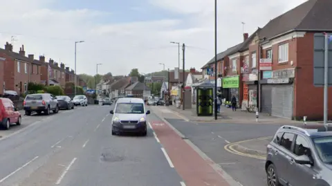 A street with cars parked on one side. Shops on one side of the street and houses on the opposite side.