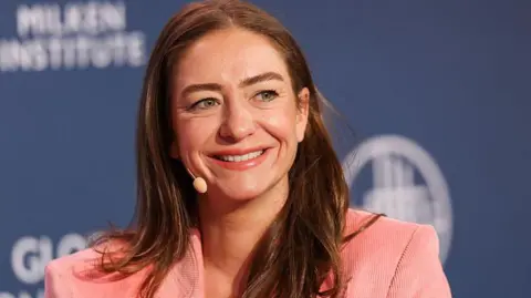Getty Images Whitney Wolfe Herd smiling while wearing a pink corduroy blazer at the Milken Institute Global Conference against a blue backdrop.