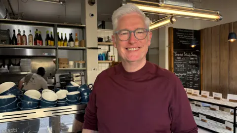 Simon Thake/BBC A man in a cafe with white hair, blue-rimmed spectacles and a burgundy top. A set of mugs are on the counter over his right shoulder. A chalkboard menu and bottles on the shelves behind him can also be seen.