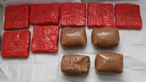 Hertfordshire Police Packages of drugs: Seven square red packages and four round brown packages lying on white paper. Each pack is marked in black with numbers or letters.