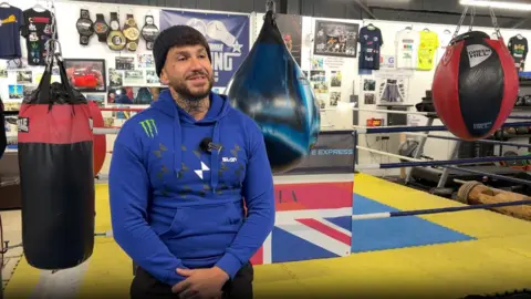 BBC Darren in a gym. He is being interviewed and wearing a mic. There is a ring behind him. Punching bags are hanging up around him. 