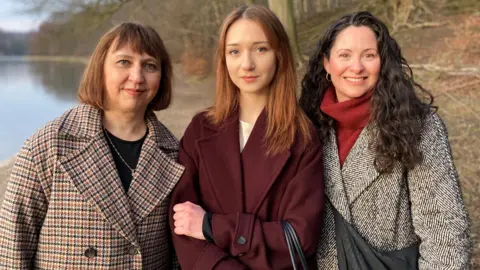 Three women wrapped up in winter clothes stand next to a lake. Barren trees can be seen in the background.