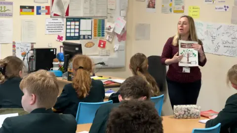 Teacher Marcella Jackson pointing to Latin posters and books in a classroom with children watching. Seven pupils, all with their backs to the camera, can be seen in the lesson.