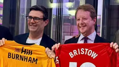 Paul Bristow Andy Burnham, left, and Paul Bristow, right, both smiling broadly and holding up football shirts bearing their names. Burnham is wearing a dark jacket over a white t-shirt and has dark rimmed glasses on. Bristow has a suit jacket, light coloured shirt and tie on.