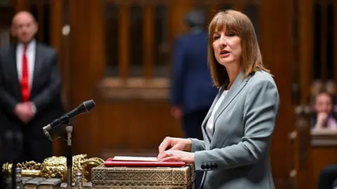 Reuters Chancellor Rachel Reeves standing at a podium in the House of Commons. She has shoulder length brown hair and is wearing a blue suit and white t-shirt. The background of the image is blurred.