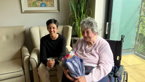 A teenage boy in a black top and beige trousers sat on a chair next to an elderly woman in a wheelchair. The woman is holding flowers and they are both smiling