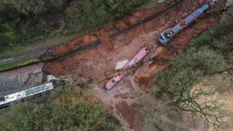 PA Media An aerial view of three boats stranded in a drained portion of a canal after a breach. 