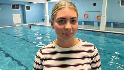 Katie Beaney is smiling at the camera. She is standing at the side of a swimming pool, where blurred images of children can be seen in the water behind her. She has fair hair, tied back, and is wearing a brown and cream striped top.