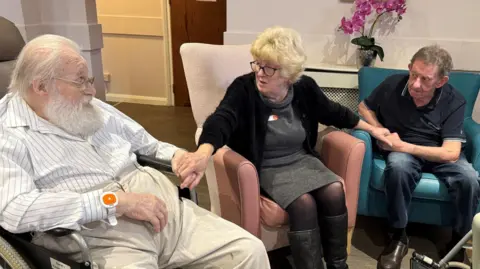 Steve Hubbard/BBC Sue Watson sits with two care home residents, who are in armchairs either side of her. She is in an armchair in the middle. She is looking at one resident as she listens to him speak. With her arms stretched out, she is holding both residents' hands. She has blonde hair and glasses and is wearing a black top, a grey dress, black tights and black boots.