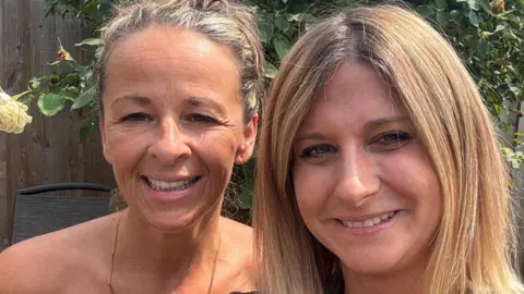 Contributed Two women smiling looking at the camera. They are standing outside, with a garden fence and trees visible behind them.