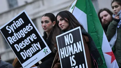 AFP via Getty Images Two female protesters hold Syrian flags and placards reading: "Syrian refugees deserve dignity" and "no human is illegal".