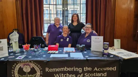 RAWS A group of four women sit behind a table - three are wearing purple tops and one is in a black dress. On the table are various leaflets and booklets. A banner draped over the table reads REMEMBERING THE ACCUSED WITCHES OF SCOTLAND.
