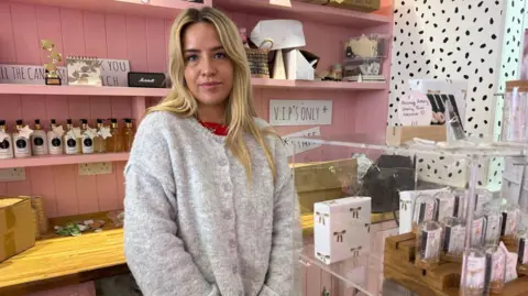 Promise Fulstow, is a shop assistant at gift shop, U Melt Me, and is pictured behind the counter next to gifts and packaging and has blonde hair and wears a wool cardigan. 