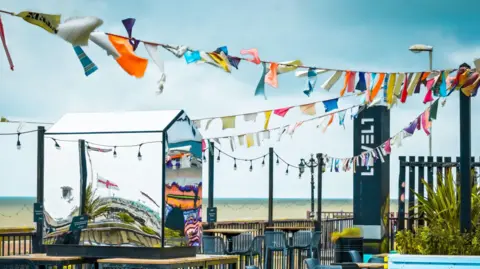 A small mirrored shed sits on a table in a pub garden. Banner decorations are strung overhead and the beach is behind.