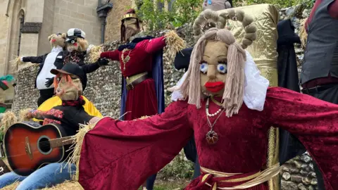 A display of themed scarecrows made from straw, each styled as a famous person, with costumes and props such as a royal dress and a guitar.