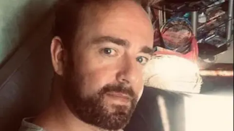 Facebook A selfie of a man on a black leather sofa with dark hair and facial hair.