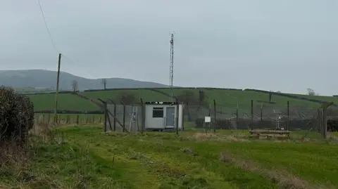 A weather station in a field. It is a small hut, with a pylon, surrounded by a fence.