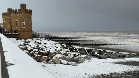 Kevin Shoesmith/BBC Two castle-like towers on a promenade, with a snow covered beach and stormy seas behind them.