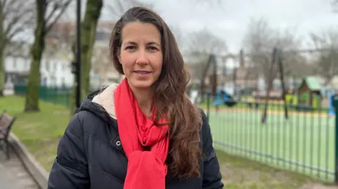 Emily is wearing a black coat and red scarf and is stood outside in front of a children's play area on a grey cloudy day.