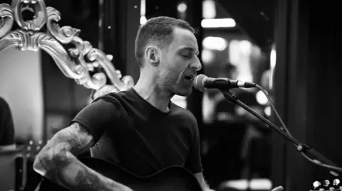 ArtHouse Jersey A black and white photo of a man singing into a microphone. There is a mirror behind him. 