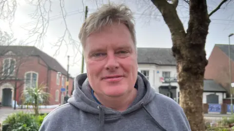 David Canty wearing a grey hoodie and standing in a town square with trees and houses in the background.