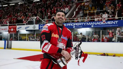Swindon Wildcats A man with dark stubble wearing a red ice hockey kit skates on a rink in front of a grandstand full of spectators. He is holding a large silver trophy