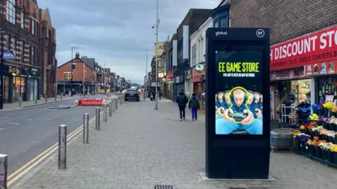 One of BT's street hub signs is in the middle of the pavement next to a discount store. It is a large black box with a screen advertising EE Game Store. People are walking down the street behind the hub.