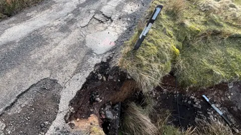 A close up shot of a narrow single track road, part of which has fallen into the river below.