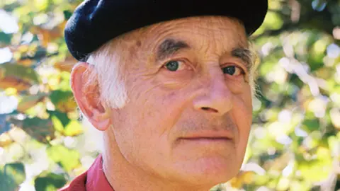 Michael Laskey, a man who is standing outside in front of some foliage of green and brown turning leaves. The sun is shining behind him through the branches and leaves. He is looking directly at the camera and smiling and is wearing a red jumper and black beret hat.